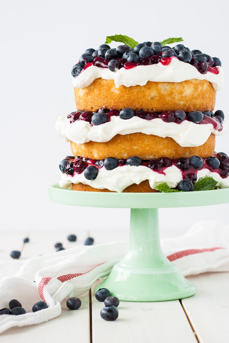 This Blueberry Shortcake Cake is the perfect light dessert for summer. Layers of vanilla cake, whipped cream, blueberry sauce, and fresh blueberries. | livforcake.com Cake shown on a mint green cake stand.