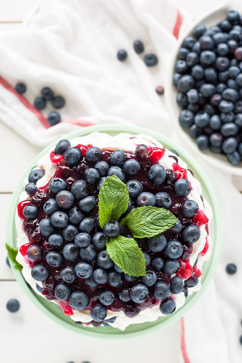 This Blueberry Shortcake Cake is the perfect light dessert for summer. Layers of vanilla cake, whipped cream, blueberry sauce, and fresh blueberries. | livforcake.com Overhead shot of the cake with the fresh blueberries piled on top. A white tea towel to the side.