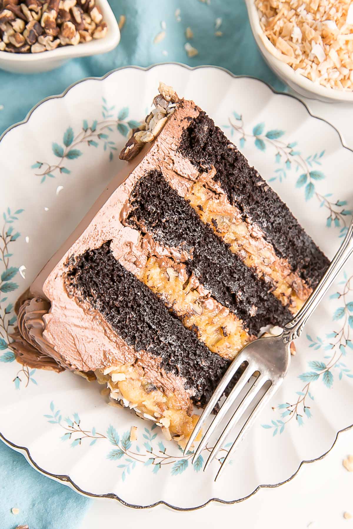 This Classic German Chocolate Cake combines rich chocolate cake layers with a sweet coconut pecan filling and a dreamy chocolate buttercream. | livforcake.com Slice of cake on a blue flower plate with a blue cloth under it.
