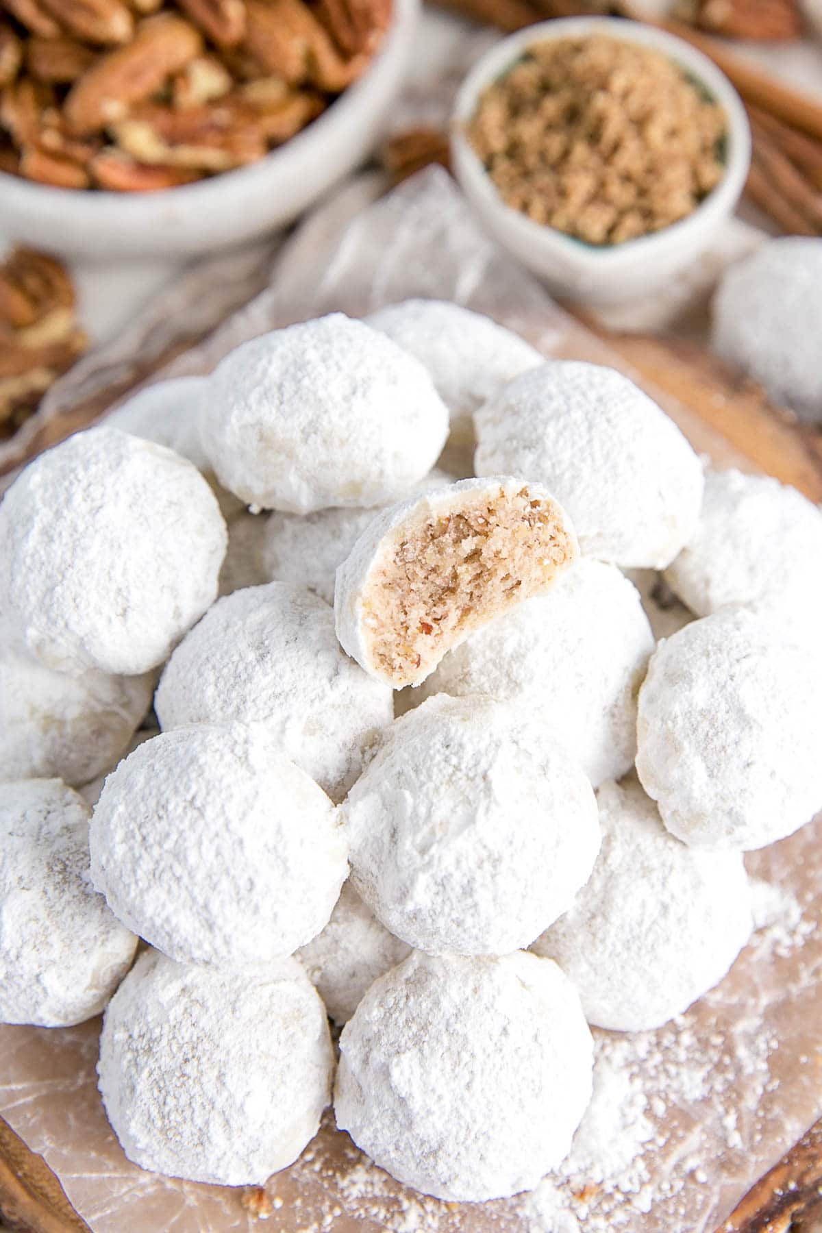 These Mexican Wedding Cookies (also known as Snowball Cookies or Russian Tea Cakes) are buttery, melt-in-your-mouth delicious! Pecan cookies with a hint of cinnamon dusted with powdered sugar. | livforcake.com Stacked cookies with pecans in the background.